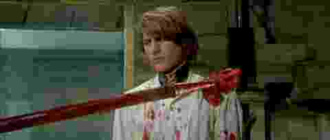 film still from the movie flesh for frankenstein. person in a bloody white shirt standing in front of a pole with an impaled heard.